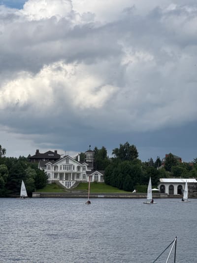 The image depicts a tranquil scene on the body of water in Chiveryovo, Russia. In the foreground, the dark blue water is gently rippled. Several small sailboats are dotted across the water, with two visible in the midground and two more further in the distance. These sailboats are white with triangular sails, and each appears to have one person on board.

In the background, a large, white multi-story building with a classical architectural style stands prominently. It features numerous windows, balconies, and a grand staircase leading up to its entrance. This building is surrounded by lush green trees and sits on a grassy embankment. To the left of this main building, a darker, older-looking structure with a pitched roof is partially visible, also nestled amongst the trees. Further to the right of the white building, another structure with arched windows and a white facade can be seen. The sky above is a dramatic mix of dark, heavy clouds and patches of brighter sky, suggesting a changeable weather day. The overall impression is one of a picturesque, somewhat grand setting by the water.