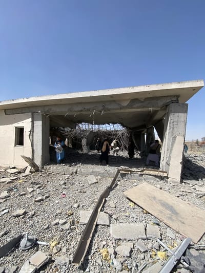 The image depicts the aftermath of significant structural damage to a concrete building in Mandisar, Afghanistan, under a clear blue sky. The main structure features a partially collapsed reinforced concrete roof, with its central section completely fallen, exposing twisted rebar. Concrete walls are fractured and show signs of severe impact or collapse.
Approximately seven to ten individuals, dressed in traditional attire, are observed moving within and immediately around the damaged structure, surveying the scene amidst the debris. One individual on the far left inside the structure wears a light blue garment and appears to be holding a white object. Another individual towards the center-right is seen crouching or bending over.
The ground in the foreground and within the building is completely covered with rubble, including shattered concrete slabs, gravel, twisted metal beams, and yellow fibrous insulation material. A large piece of plywood and a metallic object resembling a hand tamper are visible among the debris in the immediate foreground. In the background, additional rubble and distant structures or terrain are faintly visible.