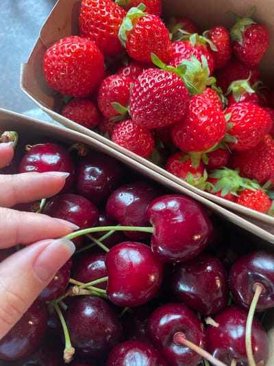 A close-up, high-angle shot shows two cardboard containers filled with fresh fruit. The container in the upper portion is brimming with bright red strawberries, some with their green stems and leaves still attached. The container in the lower portion is filled with dark red cherries, each with a slender green stem. A person's hand, with manicured fingernails, enters the frame from the left, delicately touching one of the cherries. The background is blurred, suggesting an indoor setting. The location of Sankt Anton im Montafon, Austria, is not discernible from the visual information provided.