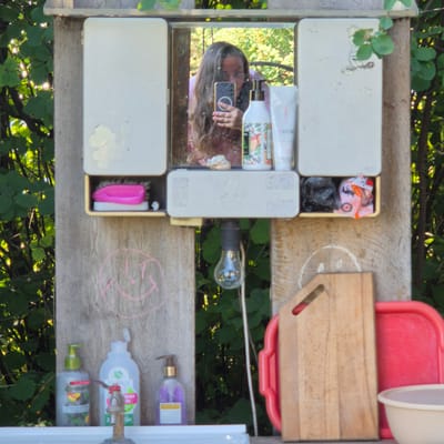 This image depicts an outdoor setup, likely in a rustic or makeshift setting, possibly in a "Novostroyka" (newly built area) in Russia. The scene is dominated by natural elements and utilitarian objects.
The main subjects are a collection of items arranged on wooden planks. A mirror reflects a person's face and upper body, holding a smartphone. Several bottles of liquid soap or lotion are visible, some with pumps. A pink brush is tucked into a small compartment. There is also a red plastic basin and a wooden cutting board. A light bulb on a cord hangs nearby, suggesting it might be an outdoor setup meant for evening use or in a shaded area.
The setting appears to be outdoors, with lush green foliage and branches in the background, indicating it's likely daytime and possibly summer or late spring, with dappled sunlight filtering through the leaves. The overall mood is casual and functional, with a hint of personal touch, as suggested by the drawing of a smiley face on the wood.
Visible text on the bottles includes brand names and product descriptions in Russian, such as " Eveline Cosmetics" and potentially other cosmetic or cleaning product labels. The text on the smartphone screen is also visible in the reflection. The arrangement of objects suggests a personal grooming or cleaning station set up outdoors.