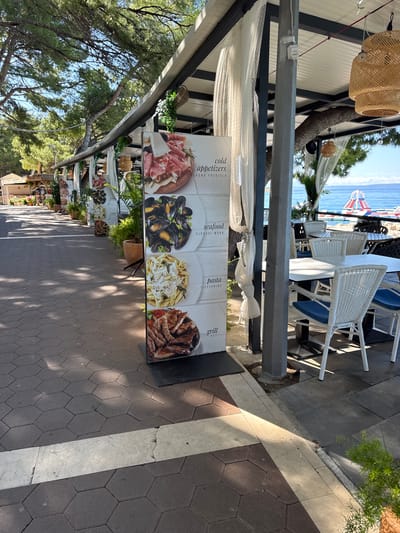 The image depicts a seaside promenade in Makarska, Croatia, with a restaurant menu board prominently displayed in the foreground. The menu board features four sections with images of food: "cold appetizers," "seafood," "pasta," and "grill." Behind the menu board, tables and chairs are set up, suggesting an outdoor dining area. In the background, the Adriatic Sea is visible, with a floating water park in the distance. The promenade is paved with hexagonal tiles, and lush green trees line the path, casting shadows on the ground. The overall scene conveys a relaxed and inviting atmosphere characteristic of a coastal resort town.