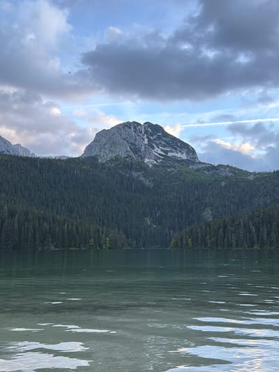 The image depicts Black Lake (Crno Jezero) within Durmitor National Park, a location in the city of Pitomine, Montenegro. The lower third of the frame is dominated by the calm, clear greenish-blue water of the lake, exhibiting gentle ripples and reflections of the sky, with lighter patches of the lake bed visible in the immediate foreground. Beyond the lake, a dense, dark green coniferous forest covers the midground, ascending the lower slopes of the mountains. In the background, rugged, grey, rocky mountains rise prominently, with the central peak featuring patches of white snow or lighter rock near its summit. A secondary, rockier peak is visible to the far left. The sky occupies the upper third, characterized by a mix of heavy grey and white clouds interspersed with patches of clear blue sky, particularly to the upper right. A distinct white contrail from an aircraft extends horizontally across the right mid-sky. Several small, dark avian figures are visible in flight against the clouds in the upper left, with one larger, darker bird or distant object present in the upper center. No human structures or explicit human activity are visible.
