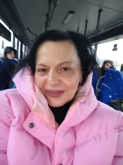A woman with dark hair and a light complexion is taking a selfie while on a bus. She wears a bright pink puffer jacket with a high collar. Other passengers are visible in the background, seated in blue patterned seats. The bus interior includes overhead lights, dark window frames, and hand rails. The overall lighting suggests an overcast day outside.