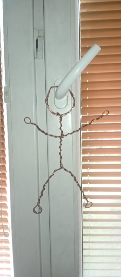 A thin, copper-colored wire figure of a person is hanging from a white window handle. The wire figure has a circular head, a twisted body, outstretched arms with looped hands, and legs with looped feet. The figure is positioned in front of a white vertical window frame and a set of light brown horizontal blinds. To the left of the window, a portion of a beaded chain or string is visible. The background suggests an indoor environment with natural light coming through the blinds. This scene is not specifically located in Karviná, Czechia.