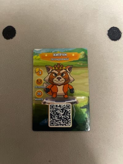 A card featuring a cartoon fox character with a crown and a star symbol is centrally displayed on a textured, light-colored surface. The card has text in Cyrillic script at the top, reading "ХИТРУН" and "ЛЕГЕНДАРНИЙ," indicating "Cunning" and "Legendary" respectively. To the left of the fox, there are circular icons with the number "3" and a cat face, followed by the text "тварини" (animals) and the number "20" with the text "номер" (number). A QR code is located at the bottom of the card. The background of the card depicts a whimsical landscape with trees and hills. Two dark circular marks, possibly ink blots or stickers, are visible on the surface to the left and right of the card. The image does not provide any information about the location in Berezhany, Ukraine.