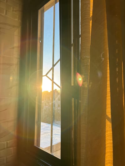 This image captures an interior perspective looking outward through a window in Mu'adammiyat ash-Sham, Syria, during either sunrise or sunset. Strong, low-angle sunlight from the exterior penetrates the window, generating prominent lens flare. The window is comprised of a dark frame, glass panes, a dark metal security grille with a diagonal and vertical bar pattern, and a fine mesh screen. To the left, a light-colored tiled wall with a subtle texture is visible. To the right, a sheer, textured yellow curtain hangs, brightly illuminated by the sun. Outside the window, a clear blue sky is visible above the horizon, where the sun appears as a bright, glowing orb. Silhouetted urban buildings are discernible in the middle distance. Below the buildings, a flat, light-colored surface, potentially a rooftop or snow-covered ground, reflects the intense light. No individuals or dynamic actions are depicted.