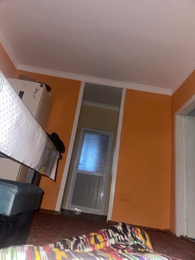 A sideways view of a room with orange walls and white trim. A window with frosted glass sits between two sections of wall. Below the window is a white appliance, possibly a refrigerator or freezer. In the foreground, a bed with a white polka dot cover and a green chair are partially visible. A colorful curtain hangs to the right side of the room.