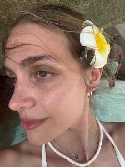 A close-up, tilted, eye-level shot depicts a woman with light brown hair and a white and yellow frangipani flower attached to her hair with a silver clip. She is wearing a white halter top and a delicate silver necklace. Her right ear is visible with a silver hoop earring.
The setting appears to be outdoors, possibly near a beach or pool, given the presence of what seems to be water in the background and a glimpse of another person lying down in the distance. The image might be taken in Lam Kaen, Thailand.