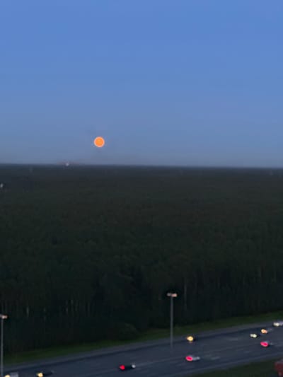 The image shows a blurry, outdoor scene at dusk or dawn. In the upper portion of the image, a bright orange circular object, likely the moon or sun, is visible against a clear blue sky. Below the sky is a dark, indistinct mass that appears to be trees or a densely forested area. In the foreground, a highway with a few moving vehicles is visible, indicated by streaks of red and white light from their headlights and taillights. There are also streetlights along the highway. The overall impression is of a landscape with a celestial body in the sky and a highway at ground level. The location is described as Imeni Sverdlova, Russia.