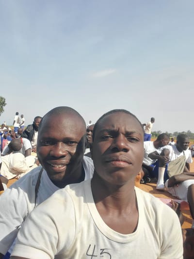 In Barakin Rafin Gora, Nigeria, two young men of African descent are the primary subjects of this self-taken photograph, positioned in the foreground. They are both wearing white t-shirts. The man on the left is smiling broadly, his mouth open, revealing teeth. The man on the right has a neutral expression. Both men appear to be outdoors during the daytime, with a clear sky in the background. The environment suggests a gathering or event, as other individuals, also appearing to be young men, are visible in the mid-ground and background. Many of these individuals are also wearing white t-shirts and are seated or standing on the ground. The ground appears to be sandy or dusty. The overall scene conveys a sense of community or group activity in an open, outdoor setting.