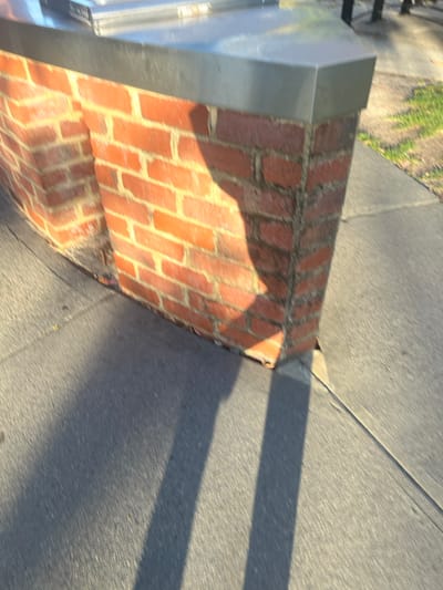 The image depicts an outdoor environment in Applecross, Australia, focusing on a section of a brick-built structure under direct sunlight. The structure features red-orange brickwork, laid in horizontal courses with grey mortar, showing minor weathering at its base. It is capped by a flat, light-grey metallic surface, consistent with stainless steel or aluminum, which forms a sharp, outward-facing corner. The ground immediately surrounding the base of the structure is a dark grey paved surface, likely asphalt or concrete, exhibiting minor surface variations and a distinct joint line. Strong sunlight illuminates portions of the brickwork and pavement, while casting a prominent, irregular dark shadow across the visible brick wall section. Additionally, long, sharply defined dark shadows extend across the paved ground, originating from the structure and off-camera elements, indicative of a low sun angle. In the blurred upper right background, faint patches of green vegetation and dark, slender vertical objects are partially visible. No individuals are present, nor are any specific actions or interactions depicted within the frame.