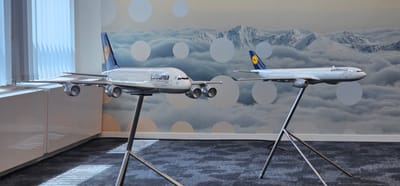 The image shows a display of two Lufthansa model airplanes. The airplane on the left is larger and appears to be an Airbus A380, with four engines. The airplane on the right is smaller and appears to be a twin-engine aircraft, possibly an Airbus A330 or A340. Both models are white with the Lufthansa logo and livery. They are mounted on silver stands and are positioned in front of a wall with a scenic backdrop of mountains and clouds. To the left of the models, there is a white cabinet with blinds covering a window. The floor is carpeted with a dark, textured pattern. The scene is well-lit, suggesting an indoor setting. The location is identified as Diegem, Belgium, likely a corporate office or public space related to aviation. There are no people or animals visible in the image. The overall impression is one of a professional display or exhibition.