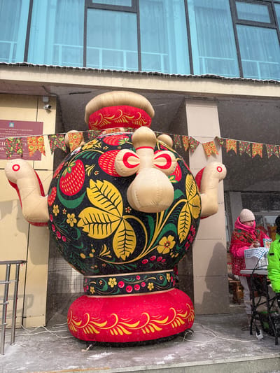 A large, inflated Matryoshka doll stands near a building with reflective windows. The doll features traditional Russian folk art designs of leaves, flowers, and strawberries. It has red and beige inflatable elements resembling headscarves and arms. Strings and banners with similar floral patterns hang nearby. The ground is covered in a light dusting of snow. A person in bright pink and green clothing is visible in the background.