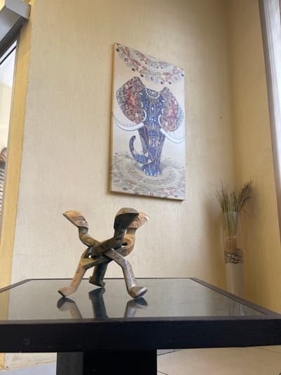 This image depicts a close-up interior shot of a room, likely in Abidjan, Ivory Coast, focusing on decorative elements. On a dark, reflective table surface, a wooden sculpture of intertwined human figures is prominently displayed. Above it, on a textured, pale yellow wall, hangs a vibrant canvas painting of an elephant adorned with intricate, colorful patterns. To the right of the painting, a tall, clear glass vase contains dried grass and is filled with small stones at the base. The wall exhibits a rough, stucco-like texture. A portion of a window with a metallic frame is visible on the left, and a white curtain can be seen on the far right, suggesting proximity to an exterior opening. The lighting suggests an indoor setting, possibly during daytime, with ambient light illuminating the scene.