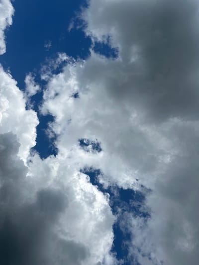 The image is an upward-facing shot of a partly cloudy sky. The clouds are a mix of bright white, fluffy cumulus formations and darker, more ominous storm clouds. Patches of deep blue sky are visible between the clouds. The scene appears to be outdoors. There are no people, structures, or any indication of location within a city. Therefore, any connection to Kinel, Russia, cannot be determined from the image alone.