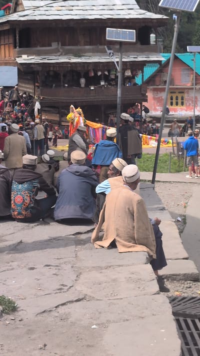 In Malana, India, a group of men are gathered outdoors, seated on a stone pathway in front of traditional wooden buildings. The architecture features multi-tiered wooden structures with ornate carvings and balconies. The men, dressed in traditional attire including caps, are observing an event or ceremony. In the background, colorful fabrics are displayed, suggesting a festive or cultural occasion. Solar panels are visible on poles, indicating a blend of traditional and modern infrastructure. The environment appears to be a village setting with cobblestone paths and surrounding buildings.