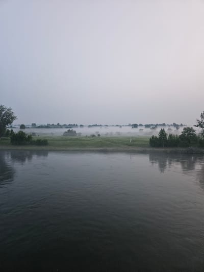 The image depicts a wide, dark body of water in the foreground, reflecting the overcast sky and the distant landscape. The water exhibits gentle ripples. In the middle ground, a grassy field is visible, partially obscured by a thick layer of mist that hugs the ground and softens the outlines of trees and low vegetation. The mist extends across the entire landscape, creating a hazy, ethereal atmosphere. The background is dominated by a pale, light gray sky, devoid of any distinct features. The scene appears to be an outdoor natural environment, possibly a riverbank or lakefront, with a rural or undeveloped setting, and it is not possible to determine the specific location within the city of Tartat, Russia from the visual information provided. There are no people, animals, or man-made structures clearly identifiable in the image, beyond what might be subtle hints of distant, indistinct objects within the fog.