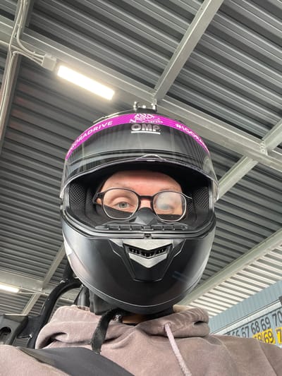 A low-angle, eye-level selfie shows a person wearing a black motorcycle helmet with a pink racing stripe and the OMP logo. The person is also wearing glasses and a black mask, with only their eyes visible. They are wearing a tan hoodie, and a black seatbelt is visible across their chest. The background consists of a corrugated metal roof and support beams, suggesting an indoor or covered outdoor structure, possibly a garage or a workshop. The image seems to have been taken at a racing or driving facility. The presence of numbers on a blue and yellow sign in the background, "67 68 69 70 69 7", is consistent with a racing pit lane or a parking area for vehicles. The location is identified as Novozhilovo, Russia.