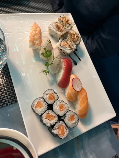 A white, rectangular plate filled with an assortment of sushi is captured in a slightly elevated, angled perspective. The plate features various types of sushi. In the upper right, there are two pieces of salmon nigiri, one adorned with a thin slice of red radish. Adjacent to these is a piece of vibrant red tuna nigiri. Below the tuna, two pieces of white fish nigiri are carefully arranged, garnished with a fresh sprig of green pea shoots. Further to the left, a single piece of pinkish-white shrimp nigiri completes the nigiri selection.

In the bottom right of the plate, six neatly cut pieces of salmon maki, featuring salmon and rice wrapped in nori, are grouped. On the upper left, four pieces of inside-out rolls (uramaki) are visible, their rice exteriors sprinkled with black sesame seeds and revealing a filling of finely chopped fish. A decorative drizzle of dark, thick sauce accompanies these rolls on the plate.

The plate rests on a dark, patterned placemat visible on the left, transitioning to a shimmering, possibly glass or polished, table surface on the right. In the blurred background, a person wearing dark attire is partially discernible. A clear drinking glass is partially seen in the bottom left corner, while a small, round dish containing a dark liquid, likely soy sauce, is cropped in the bottom right. The scene depicts a meal, possibly dinner, in an indoor setting in Basiglio, Italy. There is no visible text present in the image.