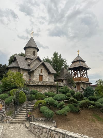 The image shows a stone church with a wooden bell tower, located on a hillside in the village of Dijelovi, Bosnia and Herzegovina. The church has a circular tower with a conical roof topped by a gold cross, and a rectangular section with a gabled roof. The bell tower is a multi-tiered wooden structure, also topped with a gold cross. The hillside is covered with manicured bushes and trees, and a stone pathway leads up to the church. The sky is overcast with grey clouds. There are no people visible in the image.