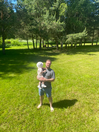 This image, captured in Копнино, Russia, displays an outdoor scene on a brightly lit, clear day, indicated by strong shadows suggesting midday or early afternoon. The foreground and midground are dominated by a lush, well-maintained green lawn or field. An adult male, appearing to be in his late 20s to mid-30s, stands centrally, facing the camera with a neutral expression. He has short dark hair and a short beard, wearing a dark grey short-sleeved t-shirt, blue denim shorts, and light-colored slip-on sandals. He holds a young child, estimated to be 1-2 years old, who is dressed in a white sun hat, a light-colored short-sleeved top, striped shorts, and light sandals. The child's back is partially towards the camera, and it faces towards the man's left. The background consists of a dense line of trees, primarily evergreens (pines) interspersed with some deciduous trees, casting shadows across the grass. A wooden bird feeder is visible hanging from a tree branch in the upper-left background, and a light-colored hammock is strung between two trees in the far-right background.