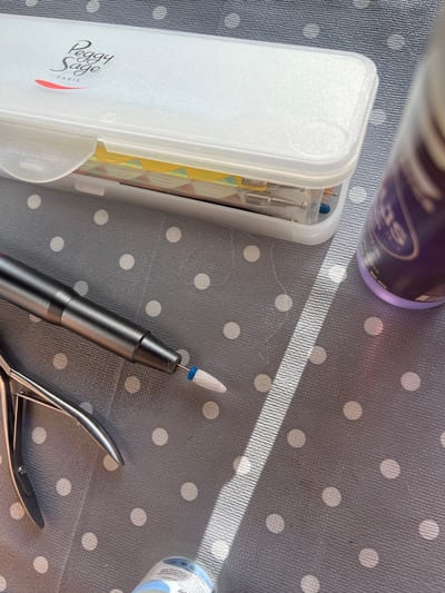 The image shows a close-up of various nail care tools laid out on a grey, polka-dotted surface. In the upper left is a clear plastic case with the "Peggy Sage Paris" logo on its frosted lid, containing an assortment of nail files or attachments. To the left of the case, a metallic nail nipper is visible. A dark grey electric nail file with a white ceramic bit is positioned centrally. To the right, a portion of a purple bottle with a metallic label is partially visible. The lighting suggests an outdoor setting with a strong shadow cast diagonally across the surface. The surface texture appears to be a textured fabric or mat. The image is taken from a top-down perspective. The scene is not indicative of any specific location in Figueres, Spain.