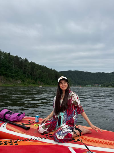 A young woman with long dark hair and a white and black baseball cap is kneeling on a paddleboard in a river. She is wearing a colorful tropical-print shirt and pants, and a light blue waterproof phone case is attached to her waist. To her left on the paddleboard is a bright pink duffel bag and a can of soda. The paddleboard is red with orange and yellow accents. The river is dark and wavy, with a densely forested hillside on the opposite bank. The sky is overcast and grey. In the distance on the river, other people can be seen on boats and paddleboards. The scene is likely in the vicinity of Манский, Russia, given the natural landscape.