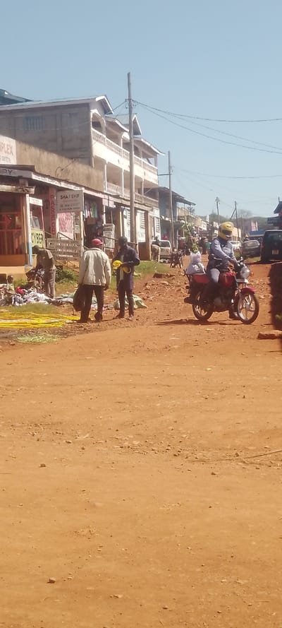 This image captures a daytime scene on an unpaved, reddish-brown dirt road in Chemuche ward, Kenya. The sky is clear and light blue.
On the left side of the road, a row of multi-story buildings with balconies houses various businesses, indicated by visible signs such as "COMPLEX," "CYBER," "PASSPORT," and "SHAJ SHIF." In front of these buildings, a street vending operation is active. A large pile of assorted clothing or textiles is spread directly on the ground. Adjacent to this, numerous long, flexible yellow objects, resembling pipes or cables, are laid out.
Three individuals are prominent on the left: one person in a dark shirt is stooping over the pile of clothing; another, wearing a light-colored jacket and a red hat, stands nearby holding a dark bag; and a third person, dressed in a dark blue shirt and black pants, stands facing the others, holding several yellow objects.
On the right side of the dirt road, a person wearing a light blue shirt and a bright yellow helmet is riding a red motorcycle. White packages are secured to the rear of the motorcycle.
Wooden utility poles with numerous overhead power lines run parallel to the street. In the background, additional buildings, people, and several vehicles, including cars and motorcycles, are visible further down the road, suggesting ongoing urban activity.