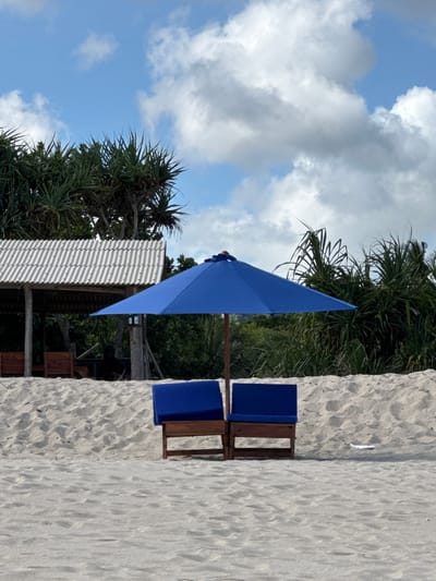 The image captures a beach scene, likely in or near Selongbelanak, Indonesia, given the tropical vegetation. A bright blue sun umbrella is positioned near two beach chairs with matching blue cushions. Behind the chairs is white sand. Behind the beach chairs and umbrella is a shelter made of corrugated metal, surrounded by lush, green tropical trees. The background reveals a partly cloudy sky with blue patches. The focus is on the umbrella and chairs, which seem to be set up for relaxation on the beach. The photograph appears to be taken during the day, suggesting a typical day at a beachside resort or location.