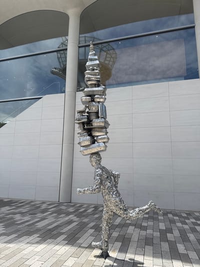 This media file shows a metal sculpture of a person in motion, outdoors. The sculpture is made of what appears to be crumpled aluminum foil, giving it a textured, almost crystalline appearance. The figure is depicted mid-stride, with one leg extended backward and the other bent forward, suggesting a running or dancing pose. The body is elongated and somewhat abstract, with the head and torso forming a single, continuous element that is stacked with various metallic, disc-like shapes.
The setting is a modern urban environment. The sculpture is placed on a walkway paved with rectangular gray and black tiles. Behind the sculpture, there is a building with a facade of large, light-colored rectangular tiles. A large section of the building features a smooth, curved, light gray architectural element. To the left of the sculpture, there is a large glass window that reflects the sky and the surrounding architecture, including a glimpse of a domed or arched structure within the building.
The time of day appears to be daytime, with sunlight casting shadows on the tiled walkway, indicating a bright and possibly sunny day. The shadows are sharp, suggesting the sun is not directly overhead. The overall mood conveyed by the sculpture and its setting is one of dynamism and modern artistry. There is no visible text in the media file. The location is identified as Aventura, United States of America.
