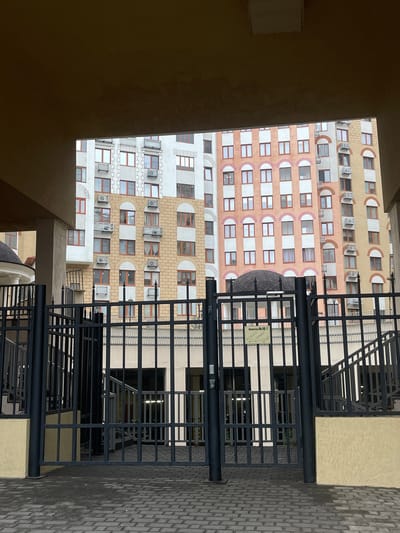 The image captures a view from an elevated position, looking out towards a multi-story building in Razvilka, Russia. The building's facade features a repetitive pattern of windows, some with air conditioning units. The exterior is adorned with a decorative motif of half-circles above the windows. A dark metal staircase and gate structure are in the foreground, partly obstructing the view of the building. The ground is paved with rectangular concrete pavers. The overall environment is urban, characterized by the dense building and constructed hardscape.