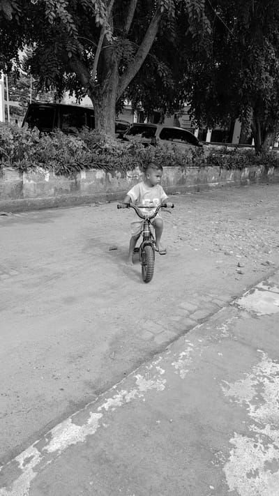In a black and white, high-angle shot, a young boy sits on a small bicycle. He is wearing a t-shirt and shorts, with his feet on the pedals. The boy is looking to his left, with a neutral expression. The bicycle is dark colored and appears to be a training bike. The boy and bike are positioned on a paved surface, which is textured and appears to be outdoors. To the left of the boy, there is a wall with some vegetation growing on it. Beyond the wall, there are trees and a car. The scene is bathed in natural light, suggesting it is daytime. The overall impression is one of a casual outdoor activity.