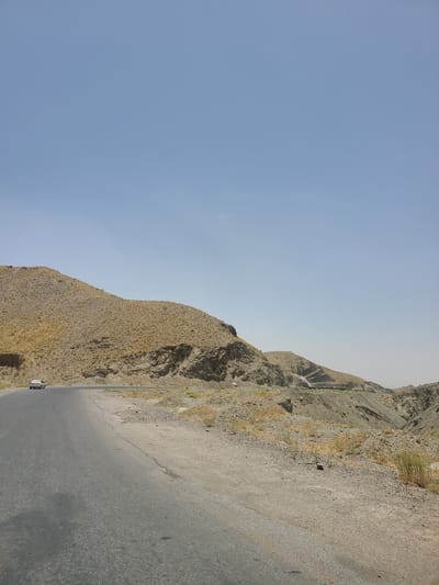 The image shows a winding asphalt road cutting through a mountainous, arid landscape. The road is dark gray, with some cracks and signs of wear. The terrain on either side of the road is steep and rocky, covered in dry, sparse vegetation. The mountains are rugged and show signs of erosion, with layers of rock and soil visible. A small white car with some red accents is visible on the road, moving away from the viewer. The sky is a clear, pale blue, suggesting fair weather. The bright sunlight casts shadows, indicating it is likely daytime. The overall scene conveys a sense of remoteness and a journey through a harsh, natural environment. There is no visible text in the image.