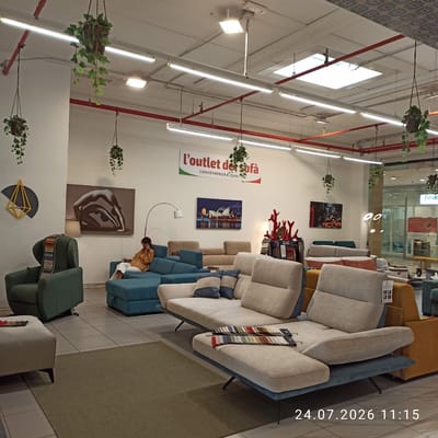 The image captures an interior view of a furniture store, specifically "L'outlet del sofà" in Modugno, Italy, as indicated by the date stamp and the store name. The scene is filled with various sofas and seating arrangements, suggesting a showroom setting. In the foreground, a large, light-colored modular sofa is prominently displayed, with fabric swatches laid out on its surface. To the left, a teal-colored sectional sofa is visible, with a person sitting on it, wearing a mustard-colored shirt and white pants, and appearing to be on their phone. Further to the left, a dark green armchair and a cream-colored ottoman are present. Several pieces of wall art are displayed, including a piece depicting two figures in a yoga pose and another featuring the Sydney Opera House. Hanging plants add a touch of greenery to the space. The lighting is a mix of fluorescent ceiling lights and track lighting. In the background, glass storefronts suggest other businesses within the same complex. The overall impression is of a well-lit, modern furniture retail environment.