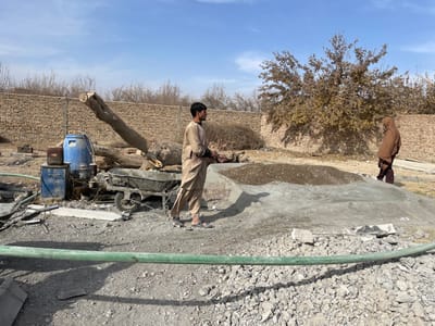 The image depicts an outdoor work scene in Nakhonay, Afghanistan, under a clear blue sky. The ground consists of gravel, dirt, and scattered debris. A light-colored brick wall with sparse, dry vegetation on top spans the background.

In the left mid-ground, a blue barrel and a piece of machinery, possibly a cement mixer or pump, are positioned. A wheelbarrow containing gray material rests beside this equipment. Several large, fallen tree trunks lie horizontally behind the machinery. A long, green hose runs across the lower foreground.

A male individual, wearing light brown traditional clothing and sandals, stands in the left-center, facing right. His hands are held at waist level. To the right, a second individual, covered in a dark brown garment that obscures their form, stands near a large, rounded pile of light gray material, topped with a smaller mound of darker aggregate. A deciduous tree with sparse brown leaves is visible behind this individual. The scene suggests ongoing construction or earthwork activities.