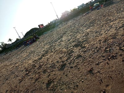 The image shows a sandy beach in Maputo, Mozambique.  Located at the coordinates -25.952 latitude and 32.614 longitude, the scene is of a mostly deserted beach. There is a single palm tree in the distance, along with a few other trees near the beach. In the foreground, there are several footprints and a few pieces of trash scattered on the sand. In the distance, there are buildings and a small group of people. The sky is clear and blue, and the sun is shining brightly.