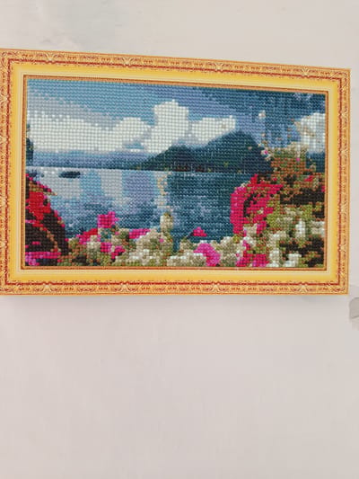 A framed diamond painting depicts a landscape scene of a lake with mountains in the background and flowers in the foreground. The painting is framed in a golden, ornate frame. The scene is composed of small, colored resin beads, typical of diamond art. The image does not provide any information to suggest it is located in the city of Койлык, Kazakhstan.