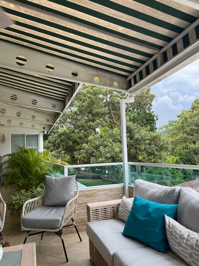 This image captures an outdoor residential patio or balcony situated in Floridablanca, Colombia, overlooking a dense, verdant natural landscape under a cloudy sky.
The patio floor is constructed of light-colored tiles. An awning overhead features alternating dark green and light-colored horizontal stripes, supported by white metal beams. These beams exhibit multiple circular perforations. A white gutter system is visible along the front edge of the awning.
The perimeter of the patio is defined by a transparent glass railing with a white top rail, mounted on a light brown or beige tiled half-wall.
Furniture present includes a light gray upholstered sectional sofa with a light-colored, possibly wicker or rattan, base. This sofa is adorned with two light gray patterned throw pillows and one solid teal throw pillow. Adjacent to the sofa is a light-colored, rectangular piece of furniture, likely a side table or ottoman, with a wicker or rattan texture. To the left, a single armchair matching the sofa's aesthetic, with a light-colored wicker or rattan frame, dark metal legs, and light gray upholstered cushions, is positioned. A partial view of an identical armchair and a glass-top coffee table is visible on the far left.
A raised planting bed, containing small green plants and a prominent palm-like plant, is situated against a white wall at the rear left of the patio. This wall also features a white sconce light fixture and a window fitted with white horizontal blinds.
The view beyond the patio railing consists primarily of a dense expanse of various green trees, forming a natural canopy. Below the main tree line, a lower green area with visible steps or a pathway is discernible. No individuals are present in the scene.