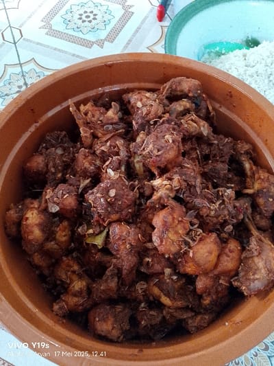 The image shows a close-up of a large brown ceramic bowl filled with a dark, rich, stewed chicken dish, likely a traditional Indonesian recipe. The chicken pieces appear tender and fall-off-the-bone, with a dark, caramelized sauce coating them. There are visible pieces of fried onion on top of the chicken, adding a contrasting texture and flavor. The dish appears to be freshly prepared and very appetizing. It is not possible to confirm the exact location in Gegesik, Indonesia, using only the coordinates provided.  The image was likely taken indoors based on the visible patterned floor tiles and the bowl's placement on a table.