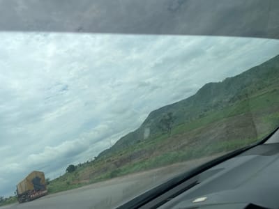 This media file depicts a scenic view from inside a vehicle, likely a car, traveling on a road in Kwali, Nigeria. The scene is dominated by a vast, cloudy sky and a lush green hillside. The road itself is visible in the foreground, with the dashboard of the vehicle partially obscuring the lower portion of the frame.
The hillside is terraced, suggesting agricultural activity. Patches of cultivated land and vegetation are interspersed across its slopes. A lone tree stands prominently on the hillside. In the distance, a large truck covered with a yellow tarp is driving on the road, its side displaying some text. The sky is filled with a mix of light and dark clouds, indicating overcast or potentially stormy weather. The overall impression is one of a journey through a rural, hilly landscape under a dramatic sky. The time of day appears to be daytime, possibly late morning or afternoon.
The text visible on the truck reads "SALAT&ANNARI".