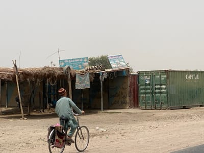 An elevated view captures a dry, unpaved roadside scene in Dand, Afghanistan, under a hazy sky. In the foreground, a male, wearing a light green tunic and a reddish-brown and beige kufi cap, rides a dark-framed bicycle away from the viewer, looking slightly left. A multicolored blanket or cloth is draped over the bicycle's rear rack. To the left, a series of ramshackle, open-fronted structures with thatched roofs and partial blue painted walls line the road, appearing as makeshift shops or stalls. A blue sign with white Arabic script is mounted above one structure, and a faded fabric sign hangs beneath it. A bicycle tire rim is visible hanging from the structure. To the right, two large, dark green shipping containers stand on the dusty ground. The container on the left displays "CAPITAL" vertically on its doors, along with the numbers "CLHU 2102636" and "22G1". The container on the right also has "CAPITAL" printed horizontally on its side. The overall environment is arid and dusty.