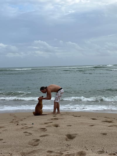 A person performs a yoga pose on a sandy beach with a dog beside them. The person is in a handstand with their body arched backward, their hands on the sand. The dog is a tan color and is looking up at the person. The beach is a light brown sand with footprints visible. The ocean is to the left of the person and dog, with waves breaking on the shore. The sky is cloudy and appears to be overcast. The scene suggests it might be daytime, possibly morning or afternoon, with the weather being cloudy. The location cues suggest a beach setting.