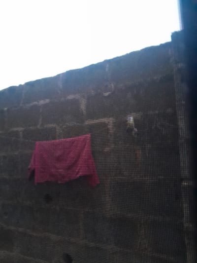 The image depicts an outdoor scene in Ifo, Nigeria, viewed through a dark, fine-mesh screen. The foreground is defined by the grid pattern of this screen. Behind the screen, a tall concrete block wall extends across the midground, featuring distinct horizontal courses of rectangular blocks. The wall's surface is dark and appears weathered, with varied textures and slight discoloration. A dark pink or maroon fabric item, possibly a towel or clothing, is draped horizontally against the lower-left section of the wall. In the upper-right portion of the wall, a small, lighter-colored object with faint yellow and green tones is partially visible. Above the wall, the sky is uniformly white, indicating bright, possibly overcast conditions. No individuals, explicit actions, or direct interactions are visible in the static scene.