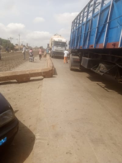 The image depicts a roadside scene in Nassarawa, Nigeria, characterized by a dusty, unpaved surface and construction work. A large blue truck with a cage-like cargo area dominates the right side of the frame, its trailer facing towards the viewer. To its left, a white Iveco truck is parked, also facing towards the viewer, with "MARIA ALLAH" written on its windshield. A person in an orange shirt and pants stands near the front of the Iveco truck. Several other individuals are visible on the left side of the image, some walking and others appearing to be involved in construction. One person in a light-colored shirt stands near a patch of what appears to be rebar and concrete reinforcing material, suggesting ongoing road or infrastructure development. A dark-colored car's headlight is visible in the lower left corner. The environment is outdoors under a cloudy sky, with a few trees and simple buildings visible in the background, consistent with a developing urban or semi-urban area. The overall impression is of a busy, functional area with vehicular traffic and construction in progress.