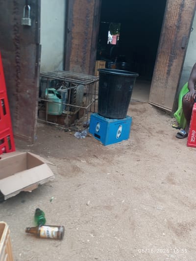 The image depicts an outdoor scene in Dokan Tofa, Nigeria, characterized by a cluttered environment with various objects scattered on the ground. In the foreground, a cardboard box lies open, and two bottles, one brown and one green, are present. A red plastic crate is partially visible on the left.

Further back, a large black plastic bin rests on a blue plastic crate. Behind these items, a metal cage structure contains a green plastic container and other indeterminate objects. The background reveals a dark doorway or opening, suggesting a building or structure. On the right side of the image, a person is seated on a green plastic chair, and a red Coca-Cola crate is visible nearby. The ground is composed of dirt and pebbles. A timestamp in the bottom right corner indicates "06/16/2026 15:55."