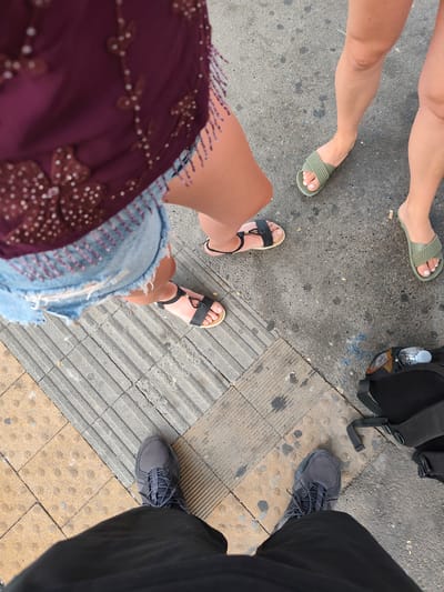 An overhead perspective shot depicts a group of people standing on a sidewalk in Carmagnola, Italy. The sidewalk is paved with square, tan-colored tiles arranged diagonally, interspersed with gray, rectangular textured tiles.
In the foreground, a person's legs clad in black trousers are visible, with a gray athletic shoe on their left foot. To the right and slightly above, a black backpack with straps is placed on the ground, partially obscuring a water bottle with a light blue cap.
Further up and to the left, two women's legs are visible. One woman is wearing denim shorts and black sandals with a braided strap. She is standing on the gray textured tiles. Another woman, to her right, is wearing a dark red, beaded top and has her legs positioned slightly above the first woman. She is wearing light green slip-on sandals. The background consists of a textured gray surface, possibly asphalt or concrete, with some dark stains.