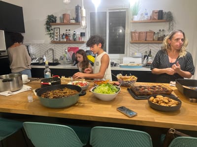 This image depicts an interior scene within a modern kitchen and dining area in a residence located in Alfey Menashe, Palestinian Territory. Four individuals are present, engaged in meal preparation or serving around a large wooden kitchen island.
On the far left, an adult woman with dark, curly hair, wearing a light-colored, off-the-shoulder top and blue jeans, stands with her back to the camera, attending to items near a stainless steel sink. To her right, a young girl with long brown hair, wearing a light-colored top, stands observing the food on the island. Next to her, a young boy with curly dark hair, wearing a light-colored sleeveless top, is actively using a utensil to interact with food in a bowl on the island. On the right side of the island, an adult woman with blonde, wavy hair, wearing a striped short-sleeved top, stands facing left, with her hands clasped in front of her and her mouth slightly open, appearing to be speaking or gesturing.
The wooden kitchen island is laden with a substantial meal. Prominently displayed are a large green pot containing a dark stew or casserole, a clear glass baking dish holding roasted vegetables and possibly meat, a dark grey bowl filled with roasted potatoes, a white bowl with mixed green salad, and another white bowl containing chopped tomato and cucumber salad. A basket with French fries and what appears to be fried fish is also present. A small shot glass containing clear liquid and a bottle of clear spirit (Absolut vodka) are visible on the island, alongside a smartphone with a reflective screen.
The background kitchen features black lower cabinets, a white chevron-patterned tile backsplash, and wooden floating shelves adorned with various bottles, jars, and potted plants. A window is visible behind the counter. The area is illuminated by recessed ceiling lights and two pendant lights. Teal/turquoise chairs are positioned at the island, indicating a dining function. The scene suggests a familial gathering centered around food.