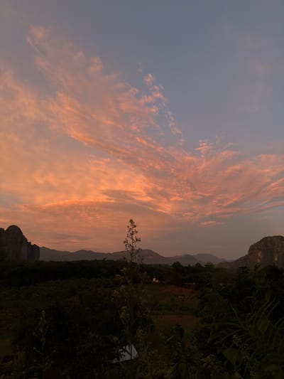 This image depicts a serene sunset scene in Vang Vieng, Laos. The sky is painted with vibrant streaks of orange and pink clouds, characteristic of a twilight sky. The landscape is dominated by lush, dark green foliage in the foreground, with a dark, imposing mountain range silhouetted against the softer hues of the sky in the background. The mountains appear rugged and distinct, suggesting a karst landscape.

The time of day is clearly sunset, indicated by the warm, diffused light and the colors in the sky. The weather appears to be clear, with the clouds being the main atmospheric feature. The overall mood is peaceful and tranquil, evoking a sense of natural beauty and the end of a day. There are no visible people or specific objects of interest beyond the natural elements of the landscape and sky. The location is identified as Vang Vieng, Laos, which aligns with the dramatic mountainous terrain and tropical vegetation often seen in that region. There is no visible text in the image.