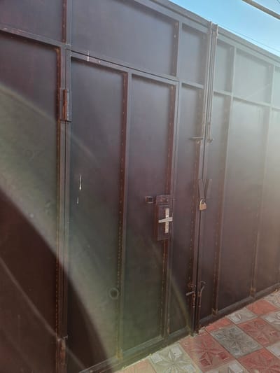 The image shows a closed, dark brown metal gate with a pedestrian door. The gate is constructed of vertical and horizontal metal bars. There are two main sections to the gate, with a pedestrian door on the left. This door has a silver-colored handle and a bolt lock. The main gate section has two vertical bolts and a padlock securing it. The gate appears to be in an outdoor setting, with a paved walkway in front of it. The paving stones are square and have a starburst pattern in red, gray, and white. The sky is visible at the top of the image, and it is a clear blue. The overall impression is of a secure entrance. The location is Saray, Azerbaijan.