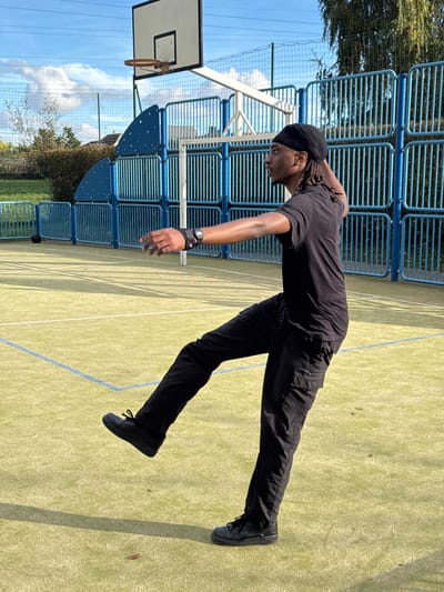 An outdoor multi-sport court in Niederhausbergen, France, is depicted under a clear blue sky with scattered clouds. A male individual, appearing to be a young adult with dark skin and dreadlocks covered by a black durag, is positioned centrally in a dynamic stance. He wears a black short-sleeved t-shirt, black cargo pants with side pockets, and black athletic shoes. His left wrist bears a black band with an integrated digital device featuring a silver buckle. The individual balances on his right leg, with his left leg extended forward and upward, bent at the knee, foot elevated off the ground. His torso is slightly rotated to the right, and his head is turned to face the right side of the frame. His left arm is extended forward with an open palm, while his right arm is bent at the elbow, hand positioned near his right ear. The court surface is light green artificial turf with faded blue demarcation lines. In the background, a white basketball backboard with a hoop and net stands to the left of the individual. A dark basketball rests on the court surface to the mid-left. The court is enclosed by a tall blue metal fence with vertical bars and curved top sections, behind which are green bushes, a lower green fence, and distant structures. Bright sunlight casts distinct shadows onto the court surface.